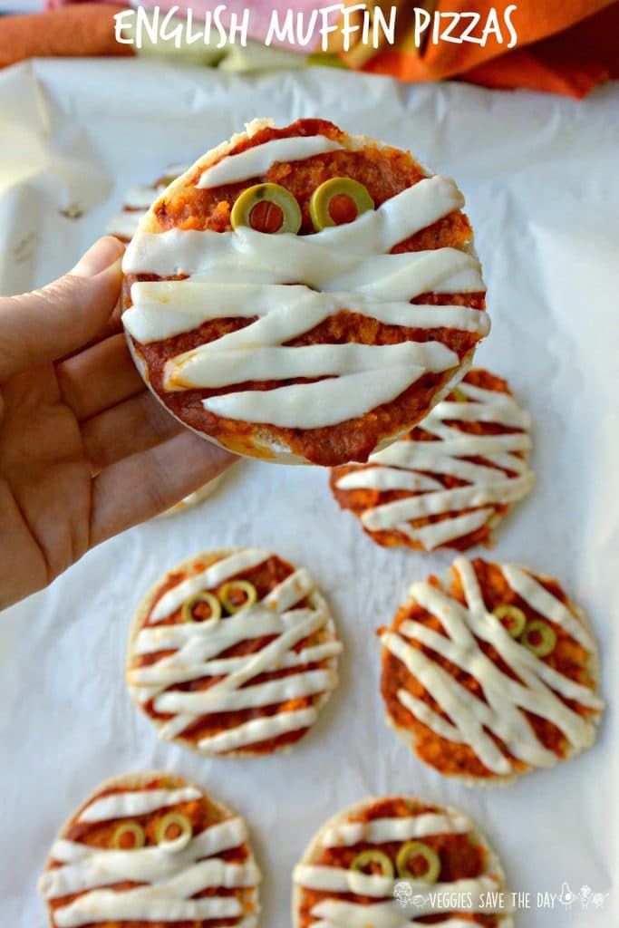 Una mano che tiene un vegan inglese muffin mummia pizza per Halloween su uno sfondo bianco.