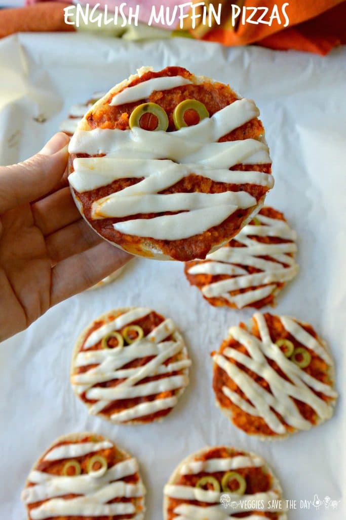 en hånd som holder en vegansk engelsk muffin mumie pizza for halloween over en hvit bakgrunn.