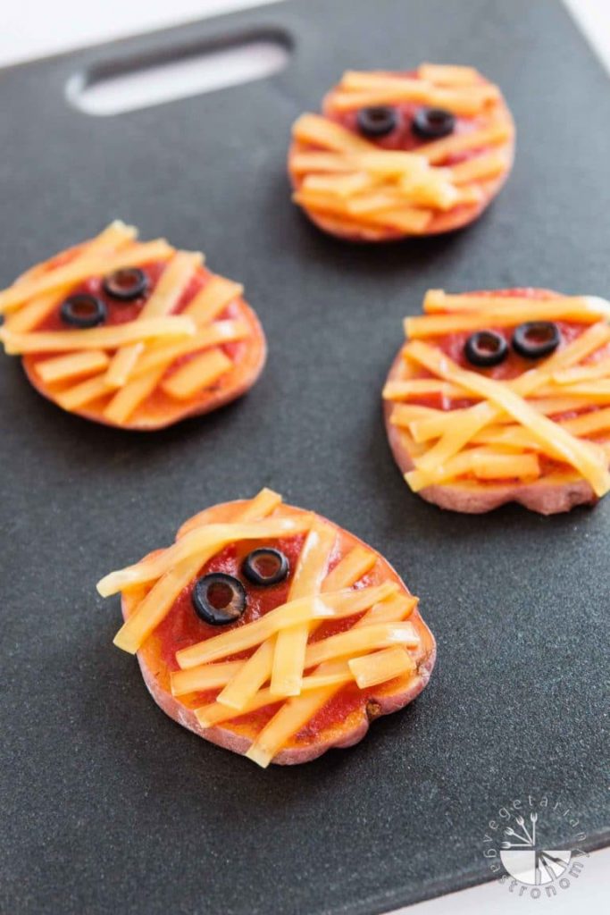 Multiple vegan mummy decorated potato slices on a dark cutting board. 