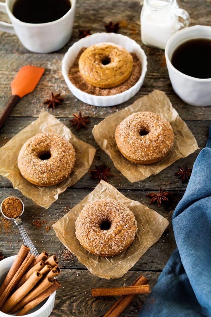 Four vegan pumpkin spice donuts on parchment paper surrounded by ingredients on a rustic background. 