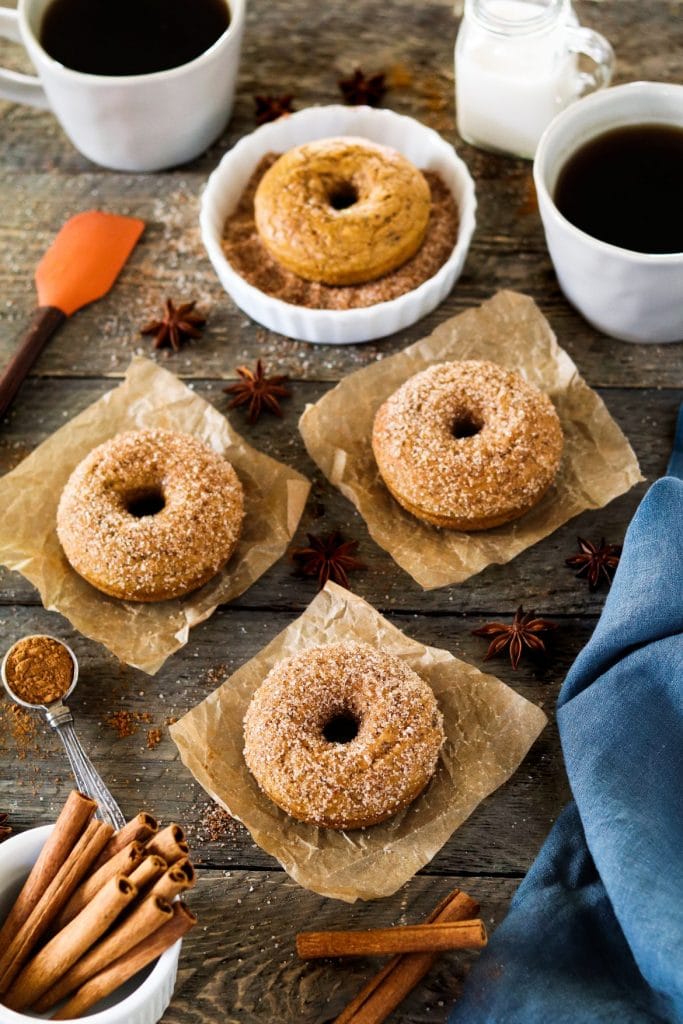  Quatre beignets aux épices à la citrouille végétaliens sur papier sulfurisé entourés d'ingrédients sur un fond rustique. 