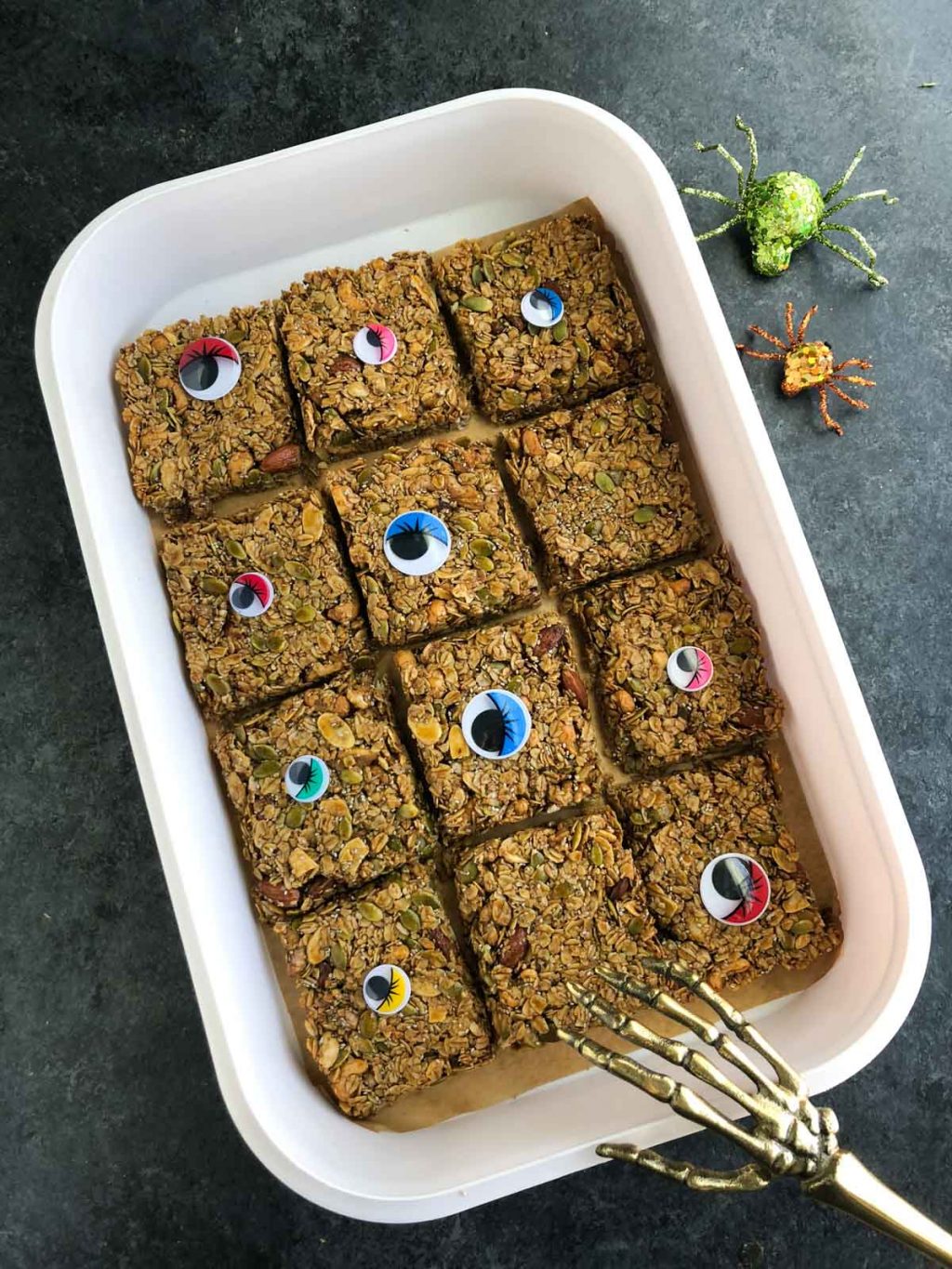 Un plato blanco lleno de barras de granola veganas junto a arañas falsas y un utensilio de esqueleto sobre un fondo oscuro.