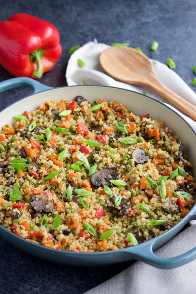 A large blue pan filled with quinoa and vegetables next to a red bell pepper and a wooden spoon on a dark background.