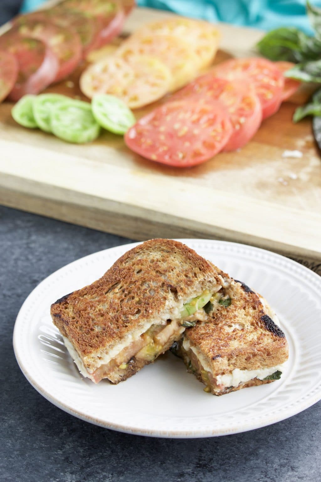 Two halves of a sandwich leaning on each other on a white plate with sliced tomatoes on a cutting board behind it.