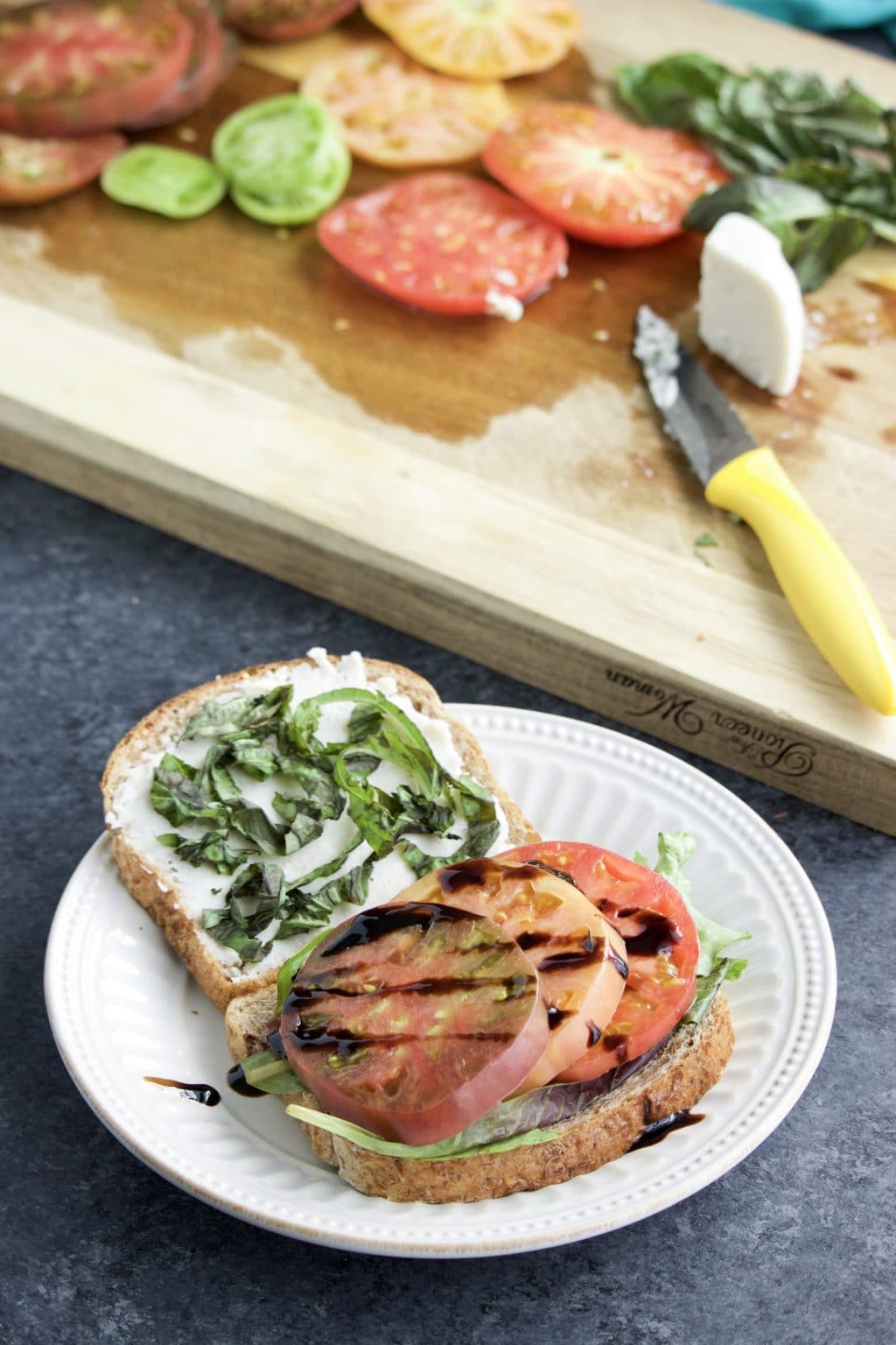 A vegan sandwich on a white plate next to a cutting board with sliced tomatoes on it. 