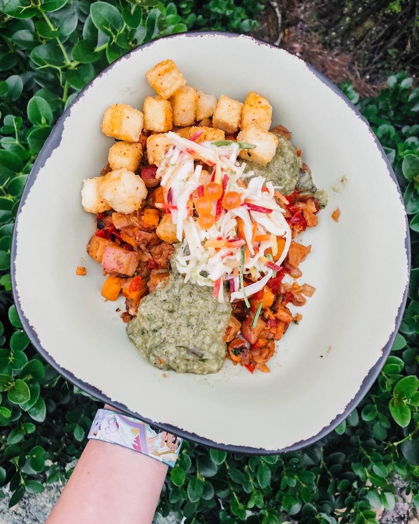 A hand holding a grey bowl that's filled with fried tofu, sweet potato hash, coleslaw, and green onion dressing over a green bush. 