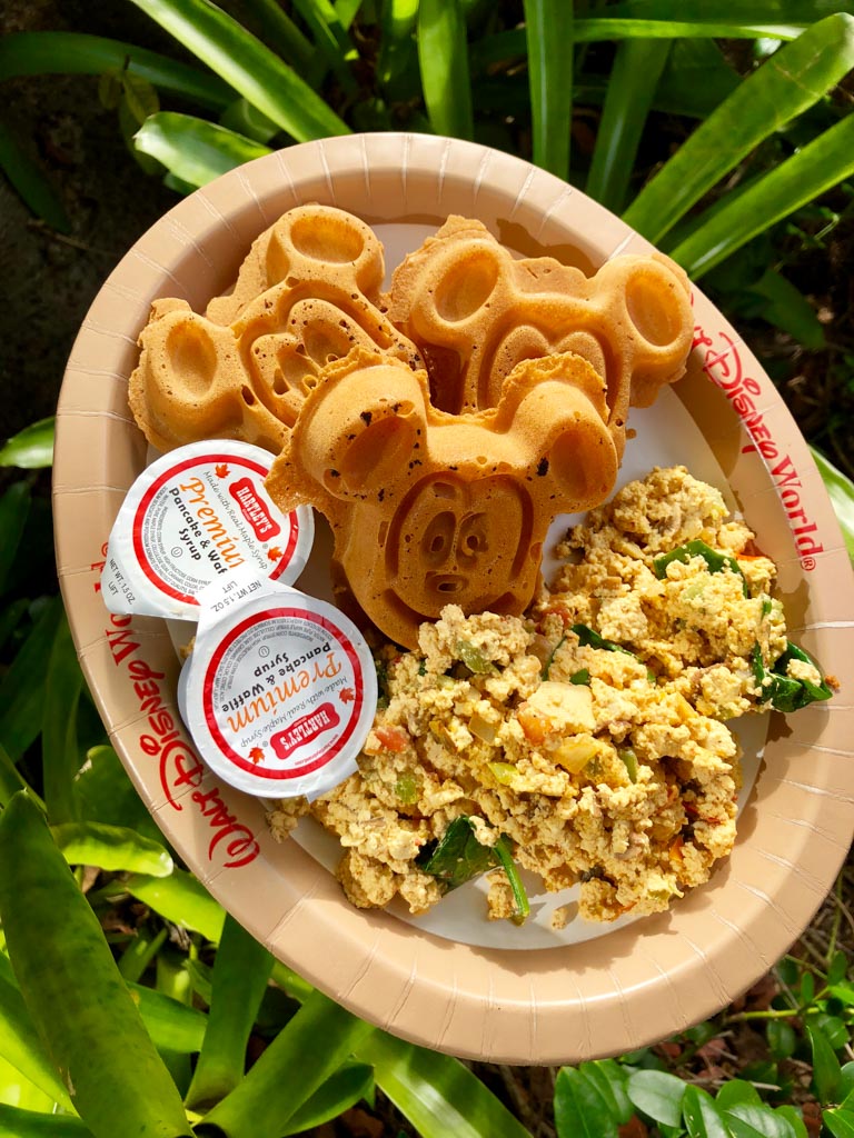 Vegan Mickey waffles with maple syrup and a tofu scramble on a Walt Disney World brown paper plate over green bushes. 