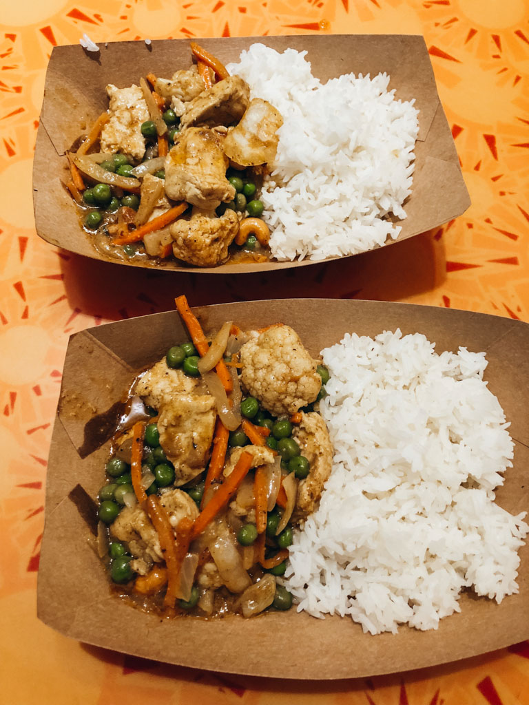 Two brown paper bowls that are filled with white rice and vegan korma on an orange table. 