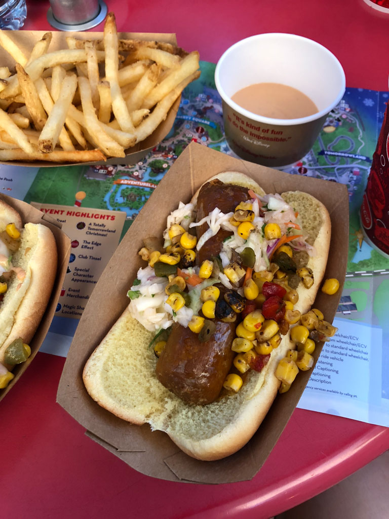 Vegan Beyond sausage hot dogs with corn salsa and French fries in brown paper cups sitting on a red table. 