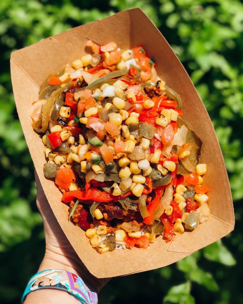 A hand holding a brown paper bowl filled with corn, peppers, onions, vegan chili, and rice over green bushes.