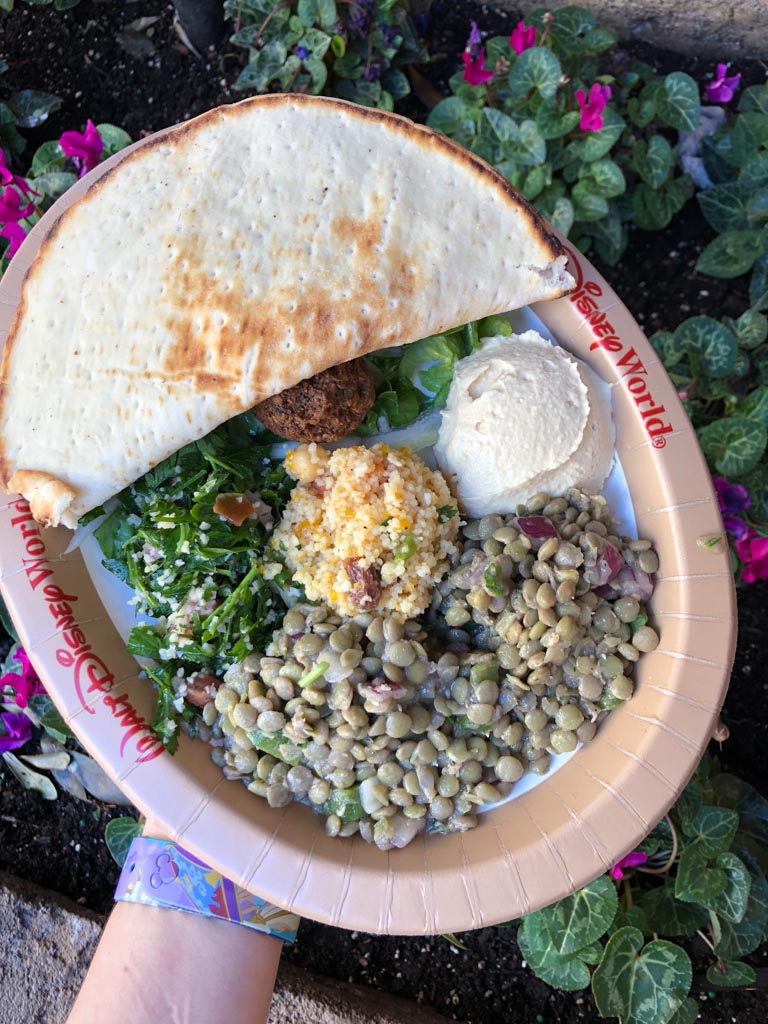 A Walt Disney World paper plate filled with vegan falafel, pita, lentil salad, tabouleh, and hummus over pink flowers. 