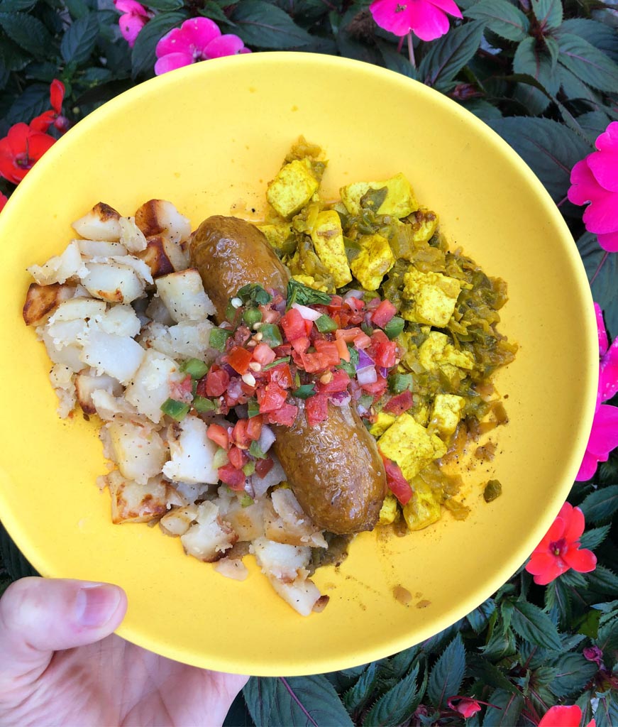 A hand holding a yellow bowl filled with vegan sausage, tofu scramble, and potato hash over flowers. 