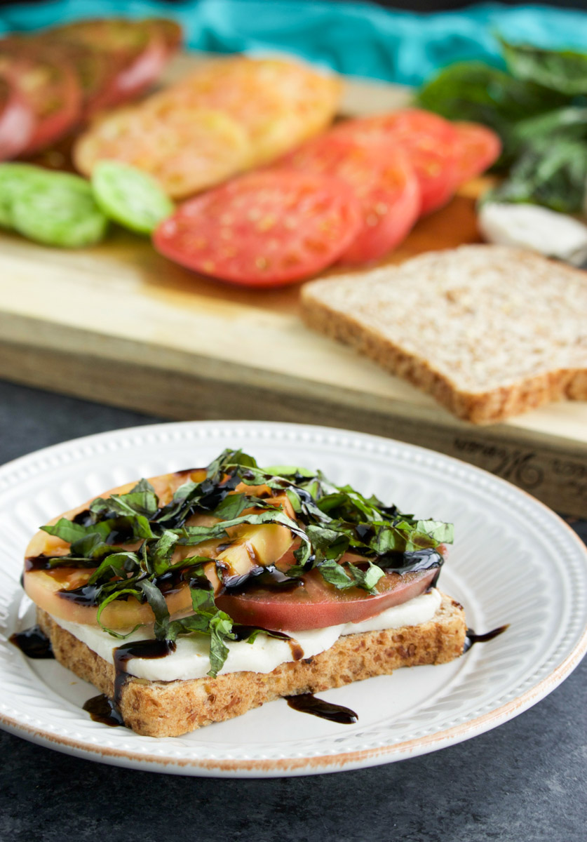 A caprese sandwich being assembled on a white plate next to a cutting board with sandwich ingredients on it.