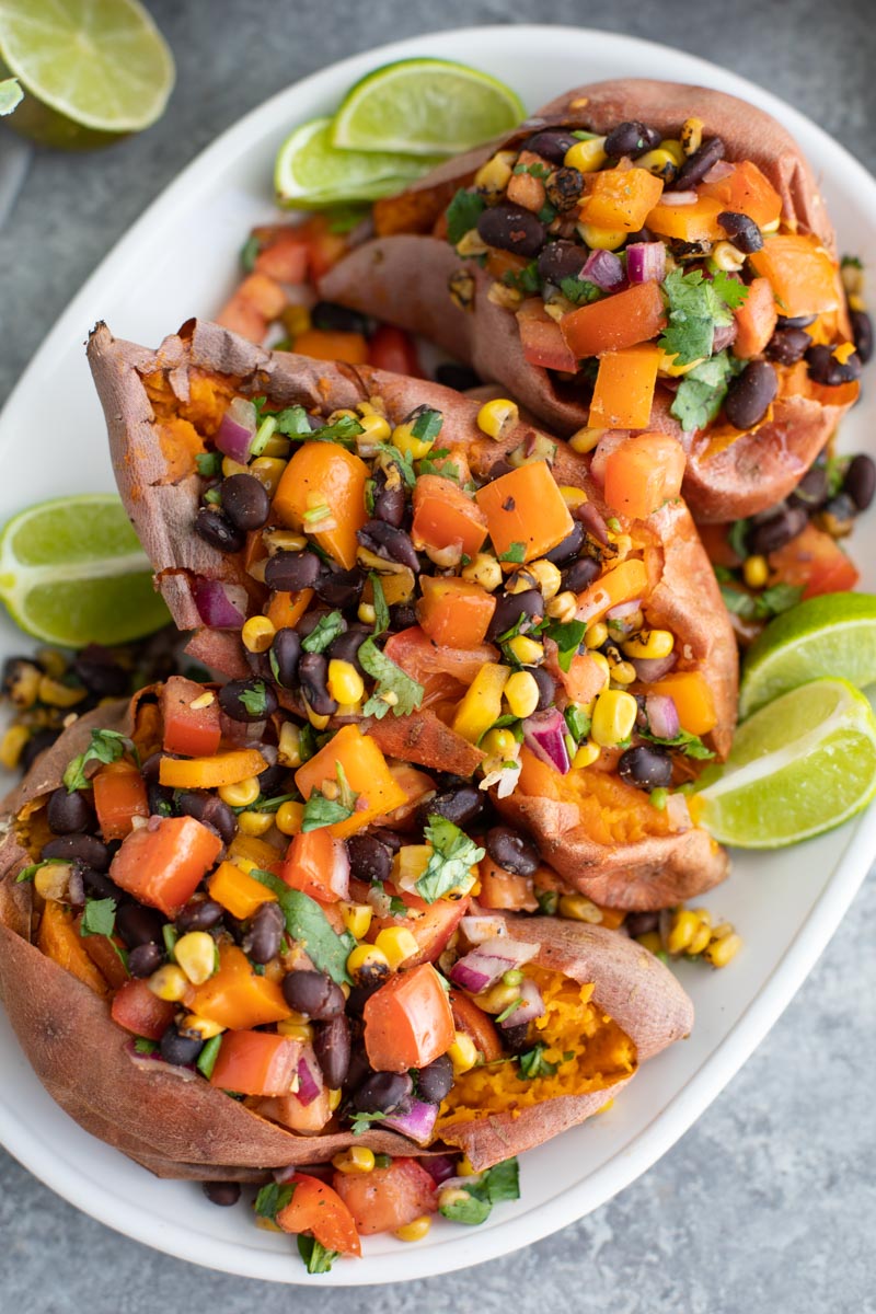 Three baked sweet potatoes on a white platter topped with vegan salsa on a gray background.