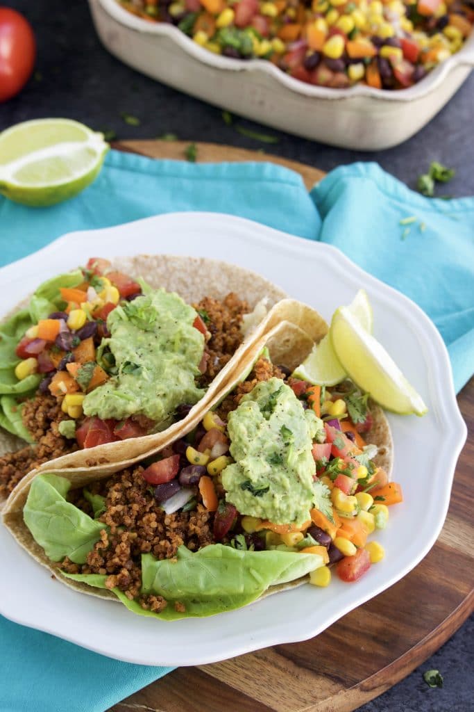 Two tacos with walnut taco meat, guacamole, and salsa on a white plate next to a blue napkin on a dark textured background. 