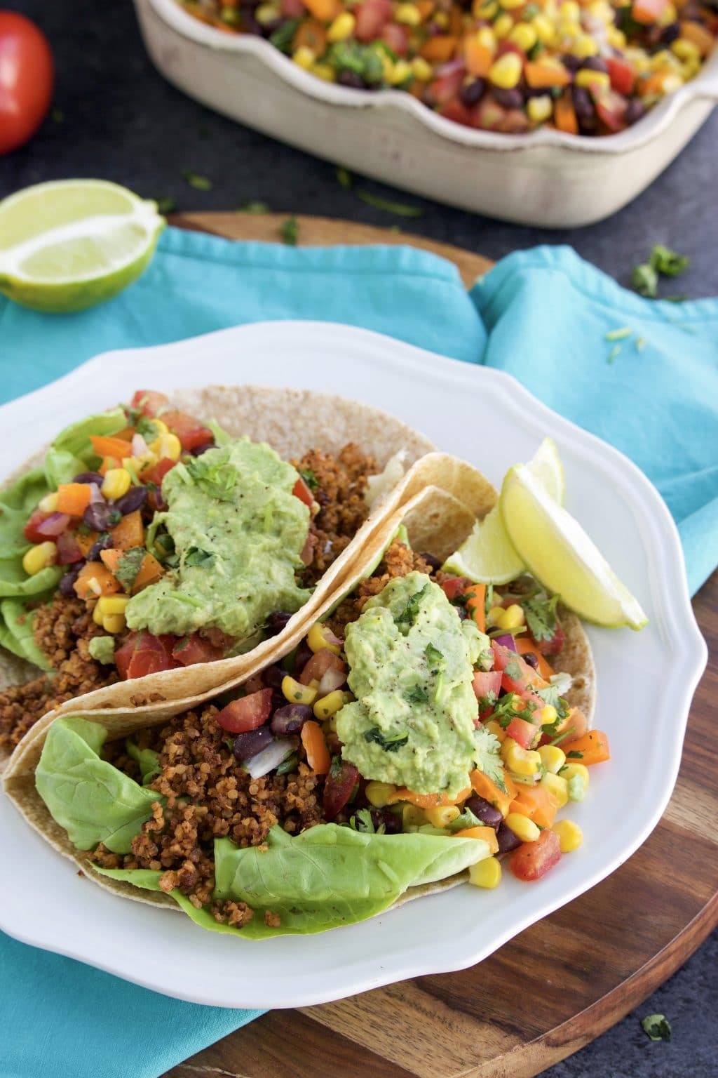 Two tacos filled with walnut meat, guacamole, and salsa on a white plate next to a blue napkin on a dark textured background. 