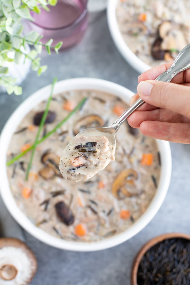 A hand holding a spoonful of soup over a filled bowl on a gray background.