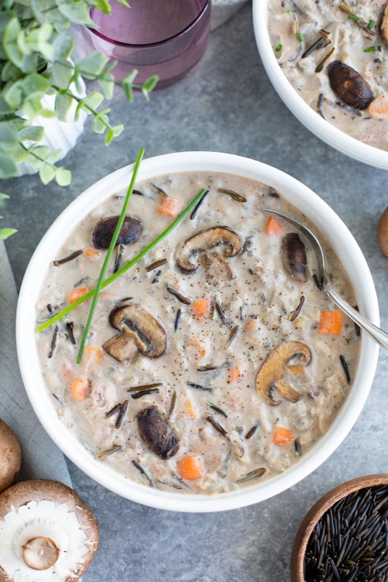 A white bowl filled with wild rice soup and a spoon next to mushrooms on a gray background.