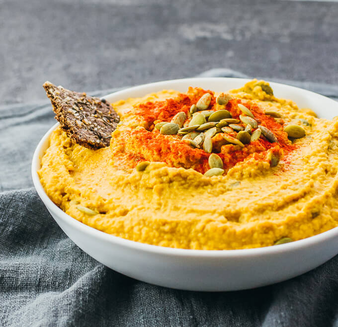 A white bowl filled with pumpkin hummus and garnished with pumpkin seeds on a dark background. 