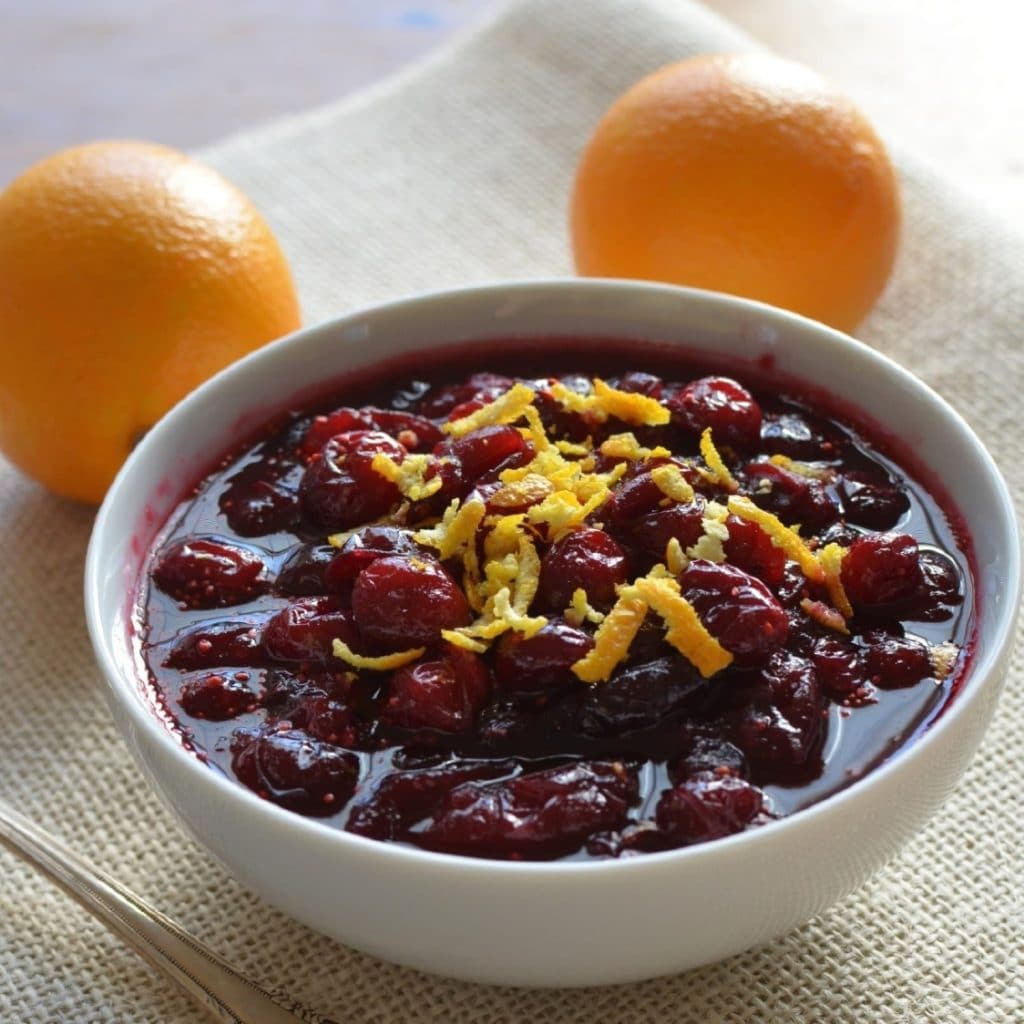 A white bowl filled with vegan cranberry sauce and garnished with orange zest next to two oranges. 