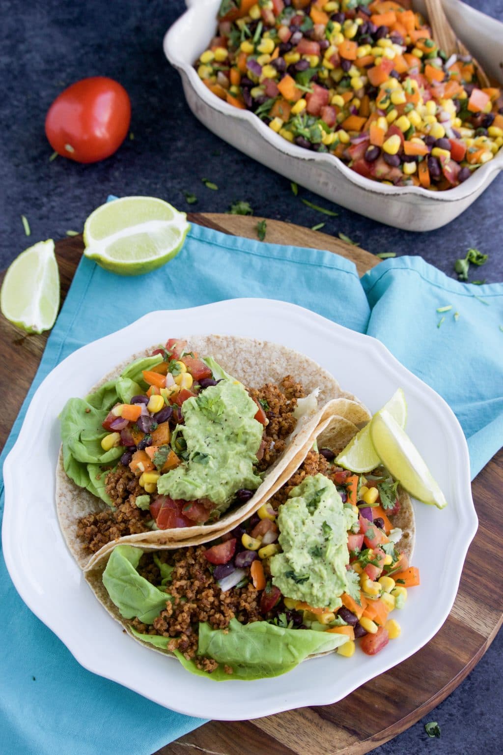 Two tacos topped with salsa and avocado on a white plate next to a blue towel on a dark textured background. 