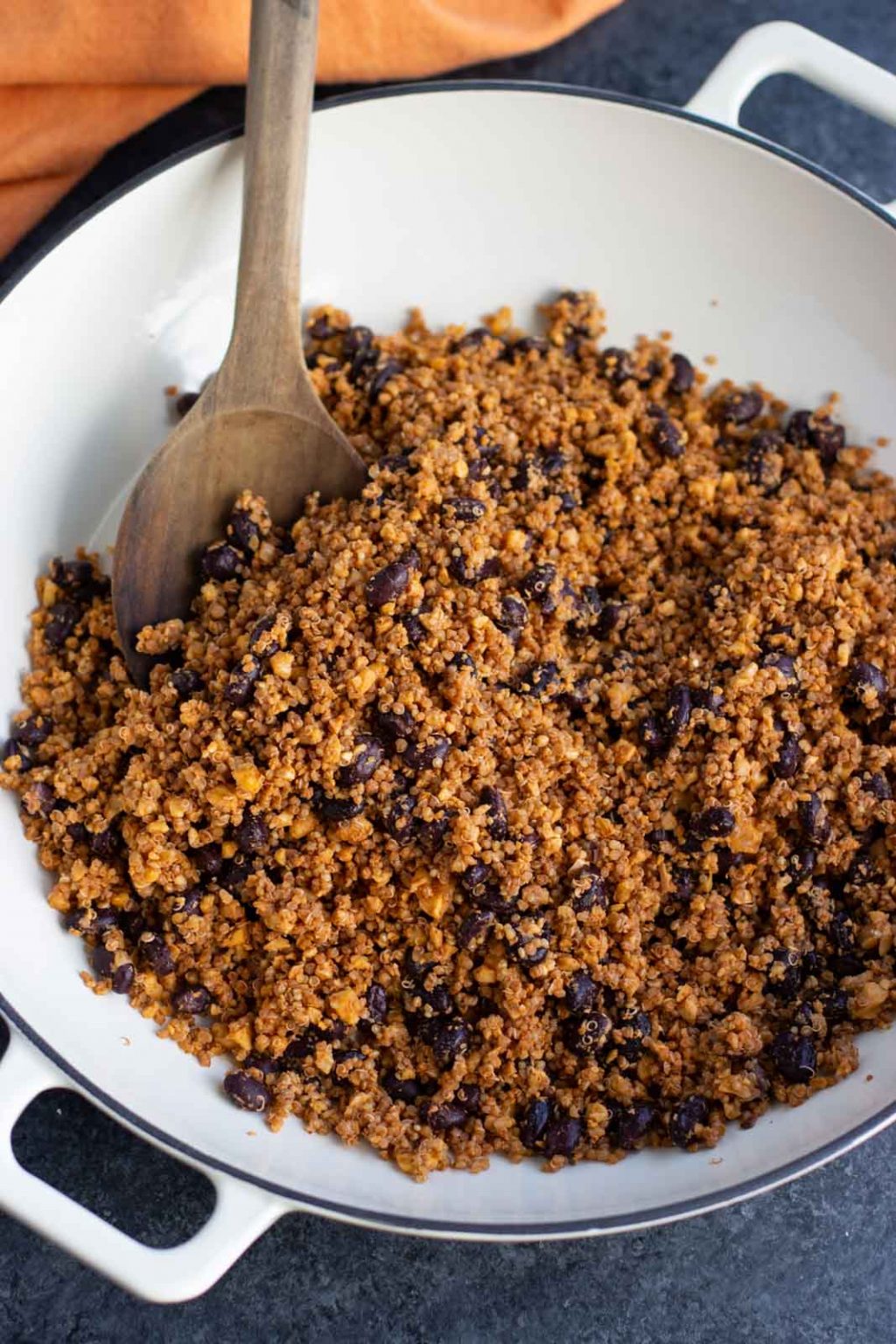 A large white pan filled with walnut meat being stirred with a wooden spoon on a dark textured background. 