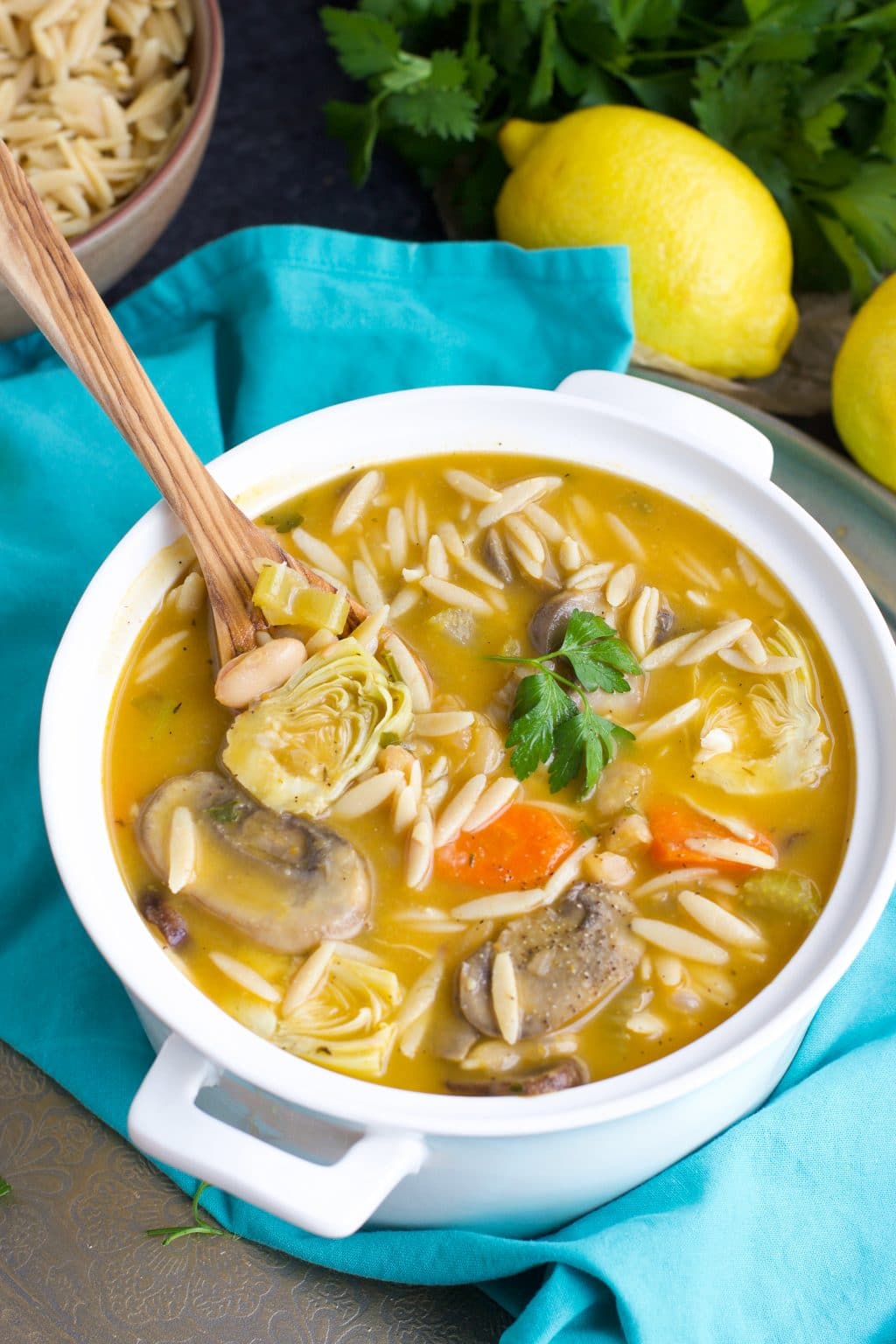 A white pot filled with soup and a wooden spoon on top of a teal towel next to lemons and parsley.