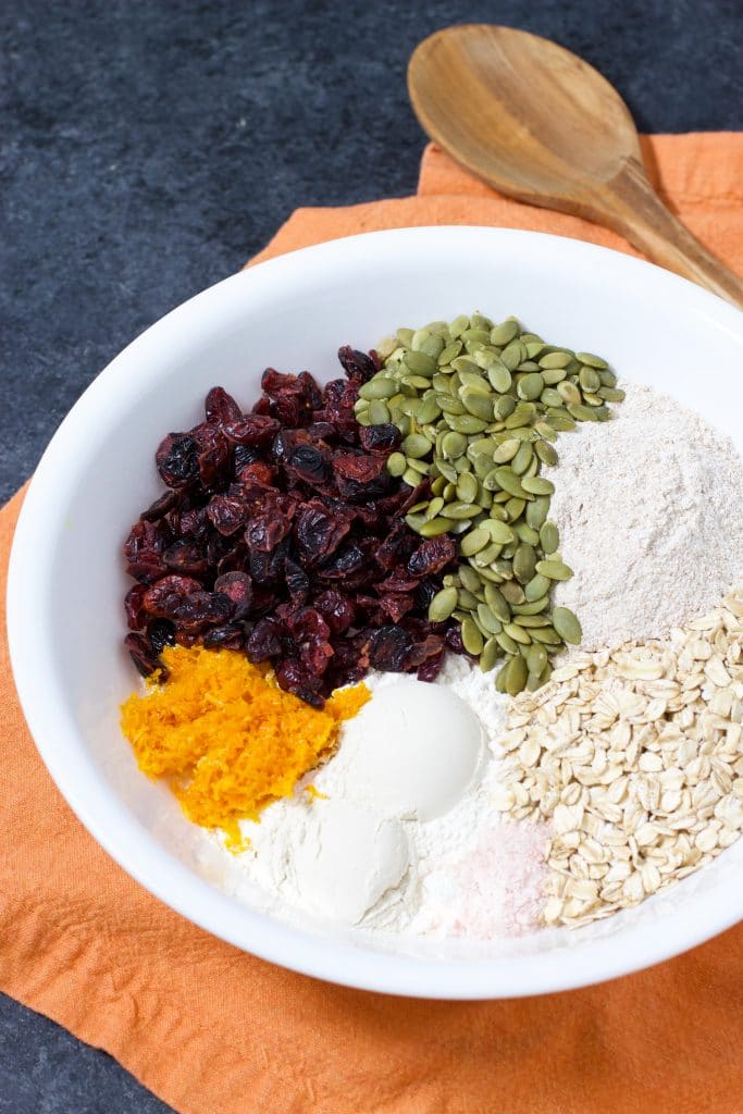 A white bowl filled with ingredients to make bread dough next to a wooden spoon on an orange napkin on a dark background. 