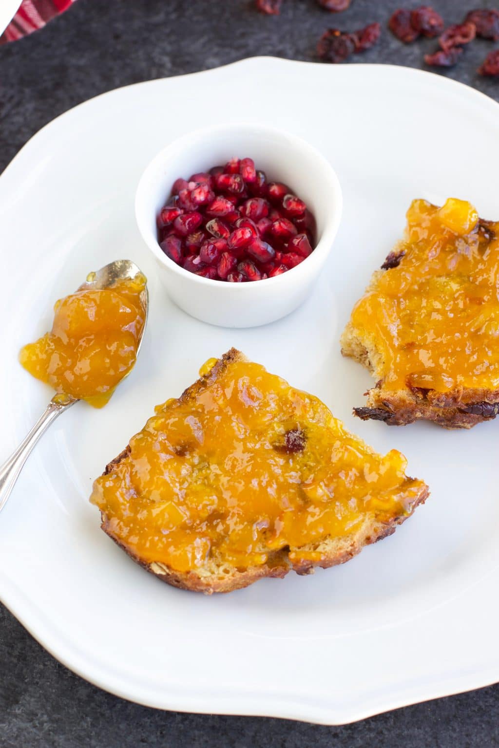 A slice of bread cut in half on top of a white plate with a small ramekin of pomegranate arils on a dark background. 