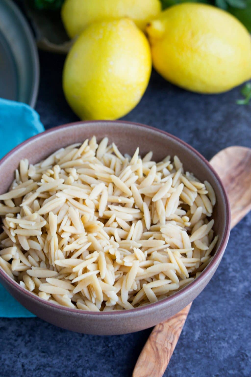 A brown bowl filled with cooked orzo next to a wooden spoon and three lemons on a dark background.