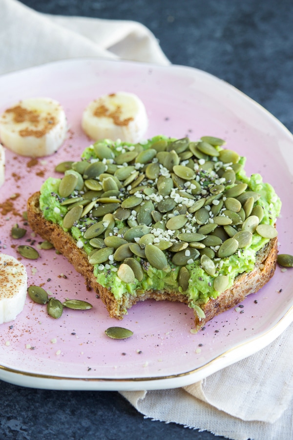 A piece of avocado toast with a bite taken out of it on top of a pink plate next to slices of banana coins on a dark background.