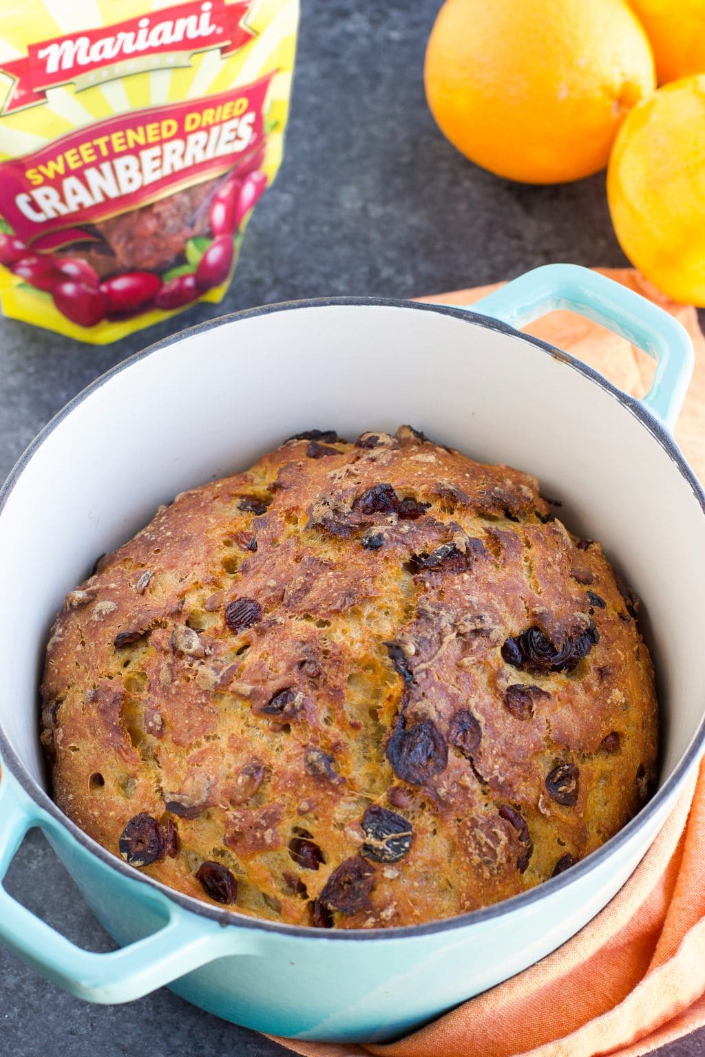 A blue cast iron pot with a load of bread in it next to a bag of cranberries and a couple of oranges. 