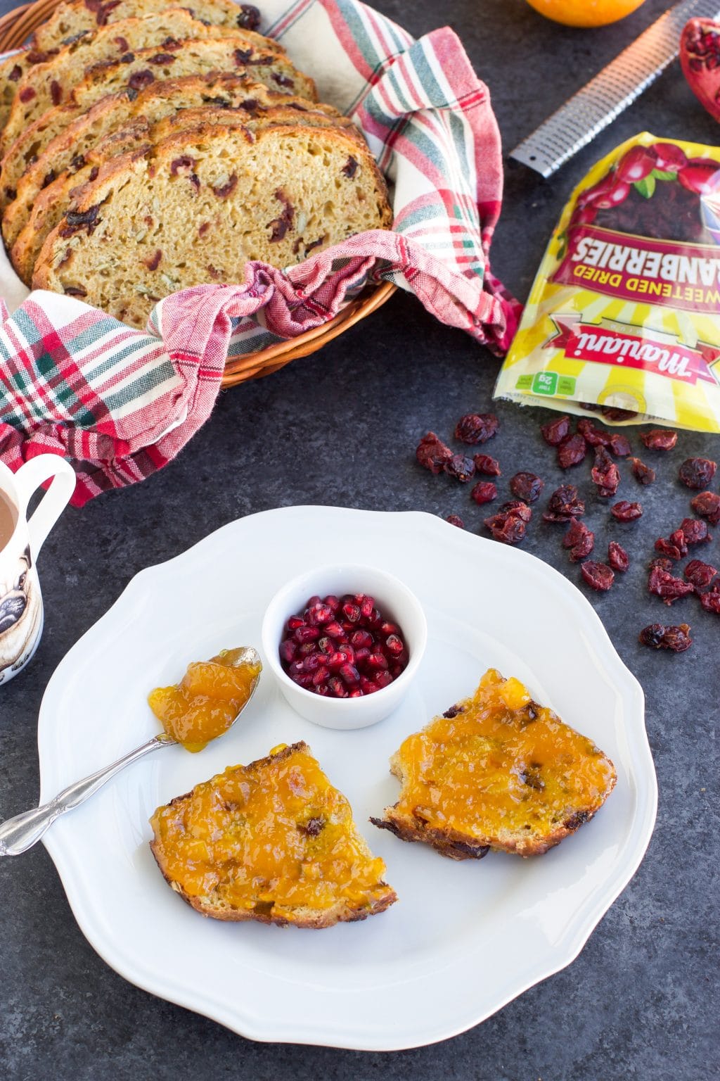 Two pieces of bread on a white plate with a small ramekin of pomegranate arils next to a basket of bread on a dark background.