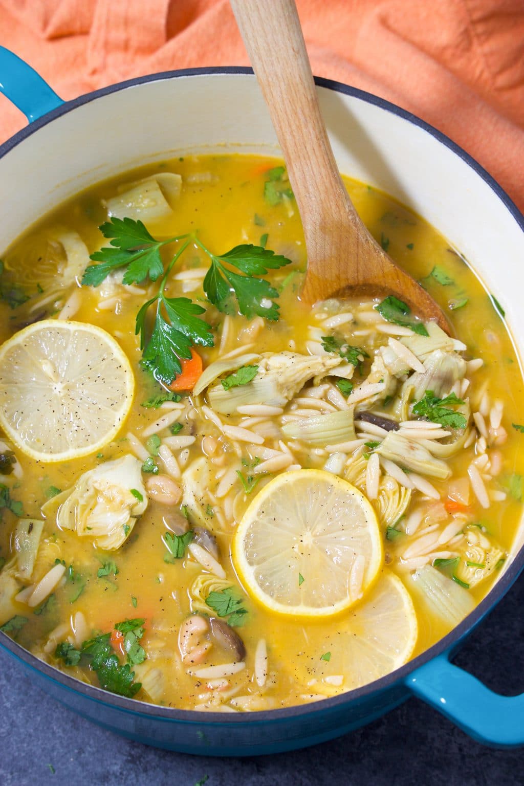 A large teal dutch oven filled with soup and a wooden spoon next to an orange napkin.