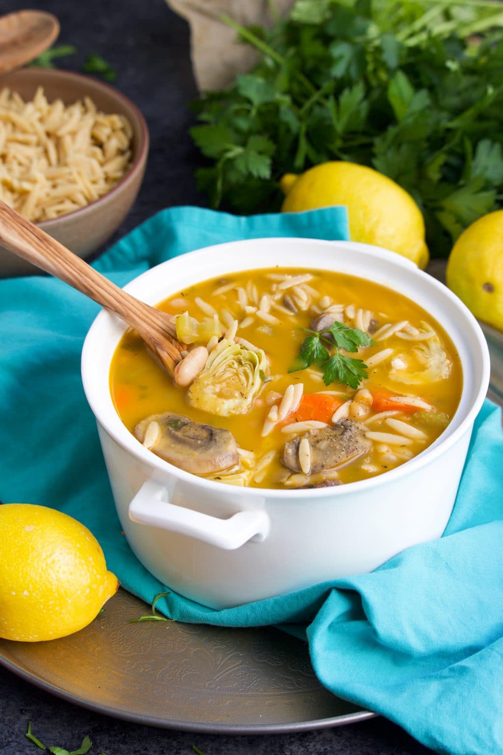 A white pot filled with soup and a wooden spoon on top of a teal napkin next to lemons and a bowl of orzo and parsley. 