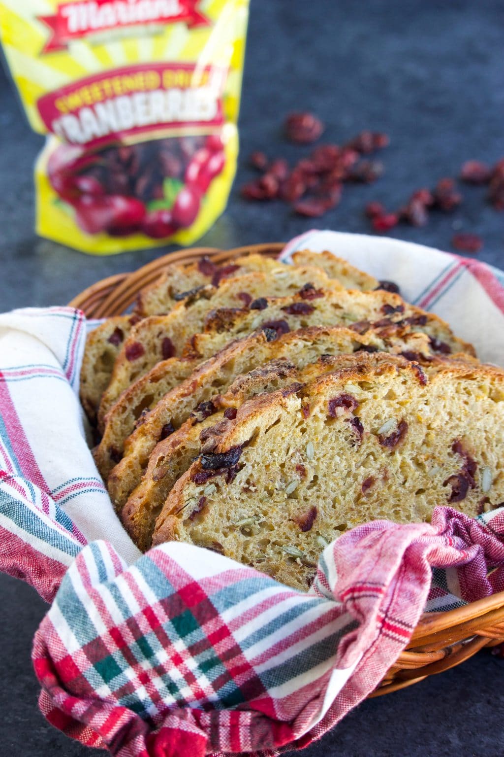 Slices of bread in a basket line with a plaid dish towel next to a yellow bag of cranberries on a dark background.