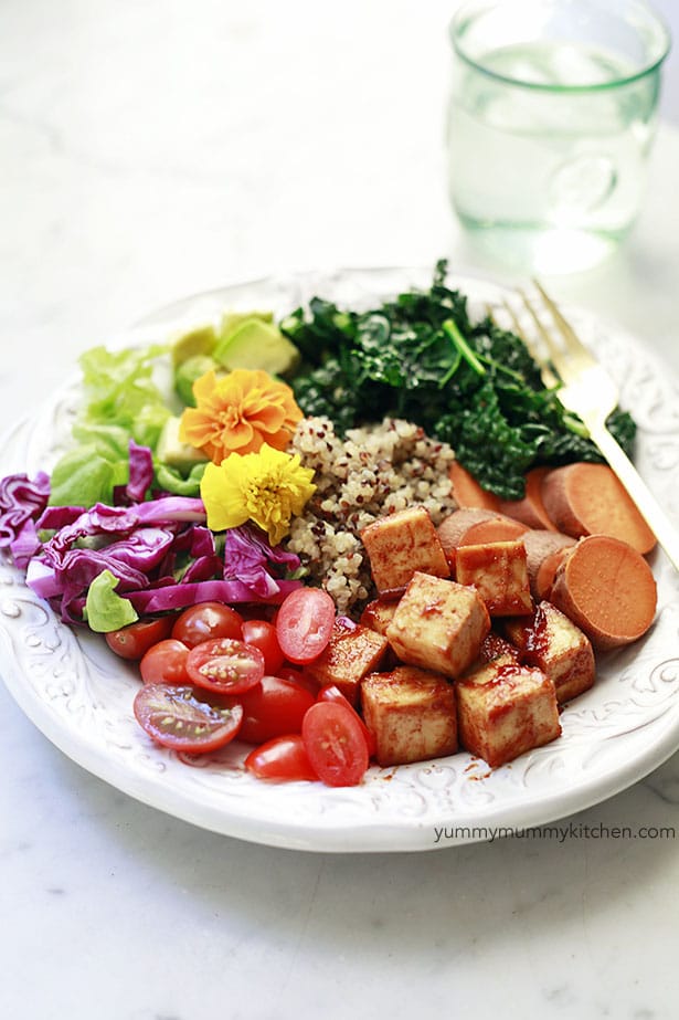 Low-carb vegan BBQ tofu with red cabbage, tomatoes, sweet potatoes, greens, flowers, and quinoa in a white bowl on a white background. 