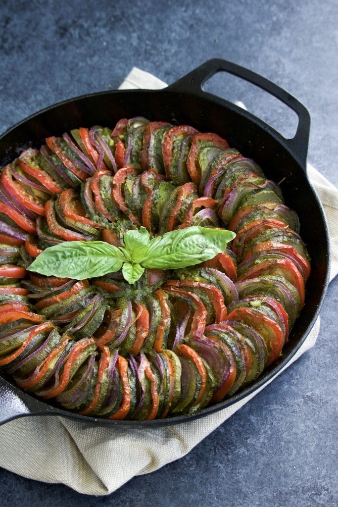 Low-carb thinly sliced zucchini, tomatoes, and red onions arranged in a circular pattern inside a cast iron pan on a textured blue background.