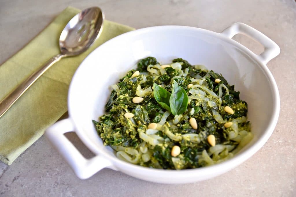 A white bowl containing low-carb vegan pesto shirataki noodles with basil and pine nuts and there is a spoon resting on a green napkin to the left. 