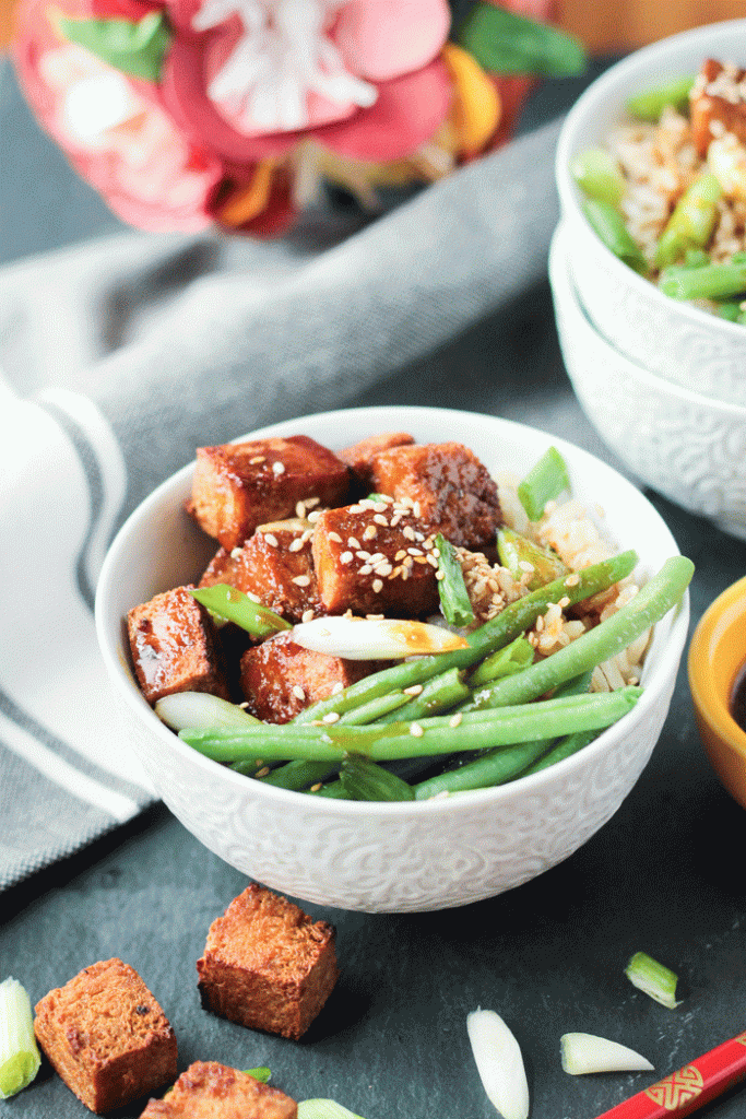 Low-carb vegan sesame ginger tofu in a white bowl with brown rice and green beans next to a grey napkin. 