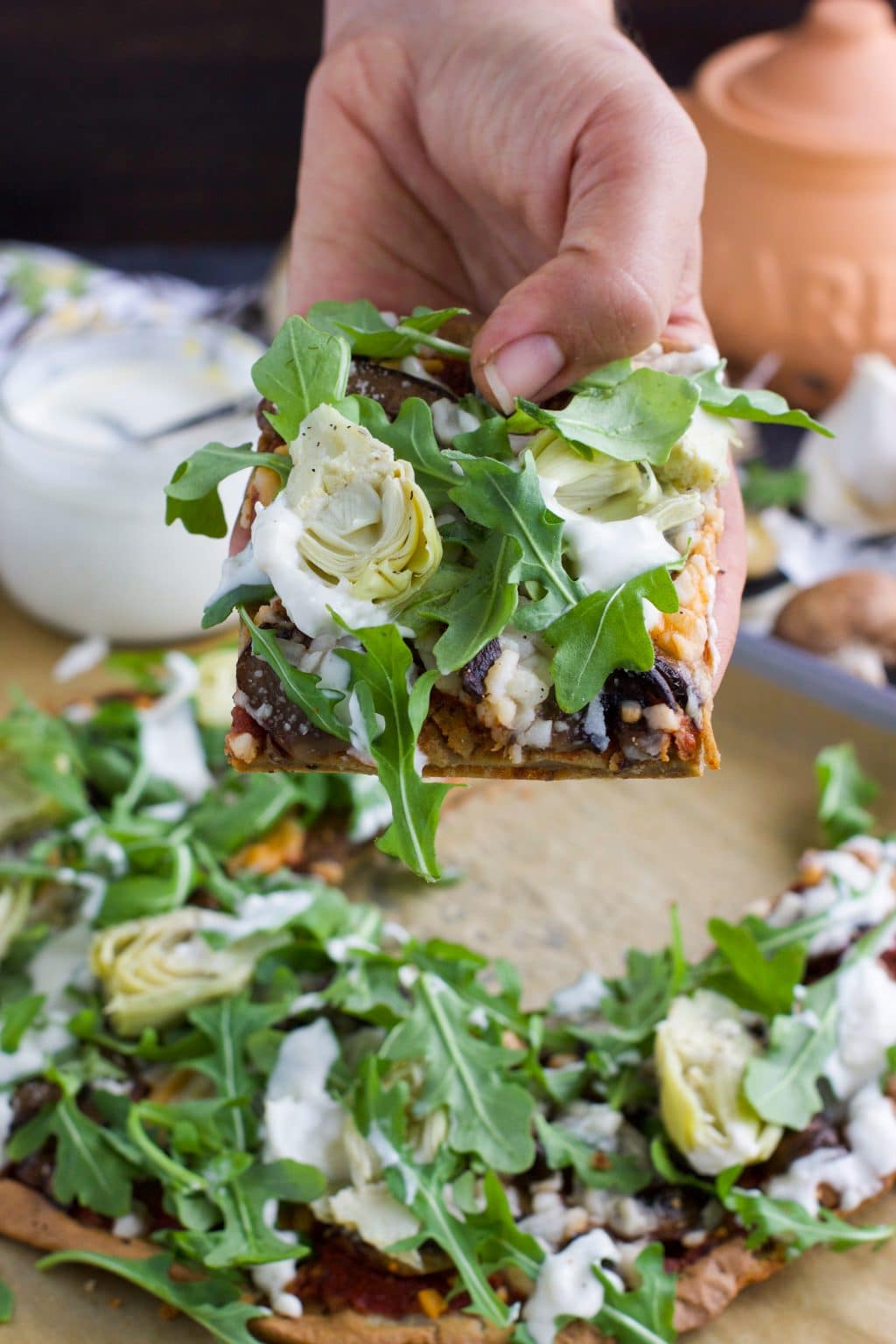 A hand holding a piece of flatbread pizza topped with artichokes and arugula.