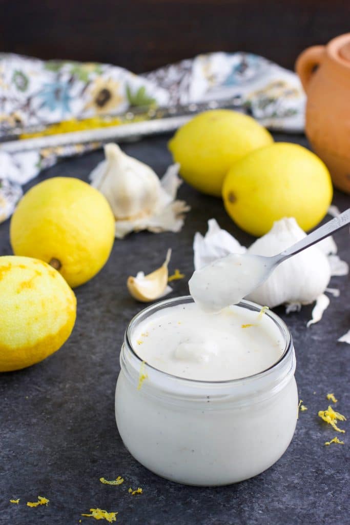 A small spoon being dipped into a glass jar of aioli with lemons and garlic cloves all around it on a dark background.