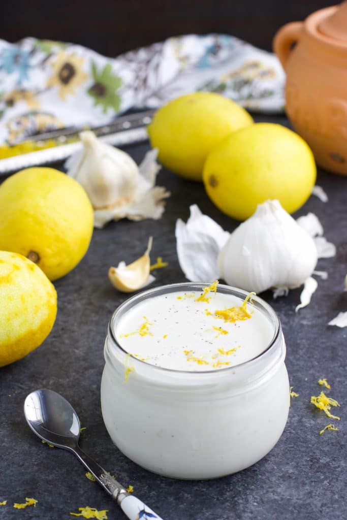 A jar of vegan keto creamy garlic aioli topped with lemon zest and it has a spoon, lemons, and garlic cloves all around it on a textured background.