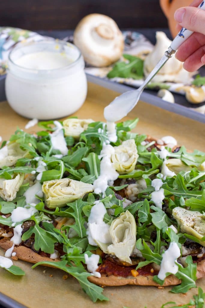 A hand holding a spoon and drizzling white sauce over a pizza on a parchment-lined tray.