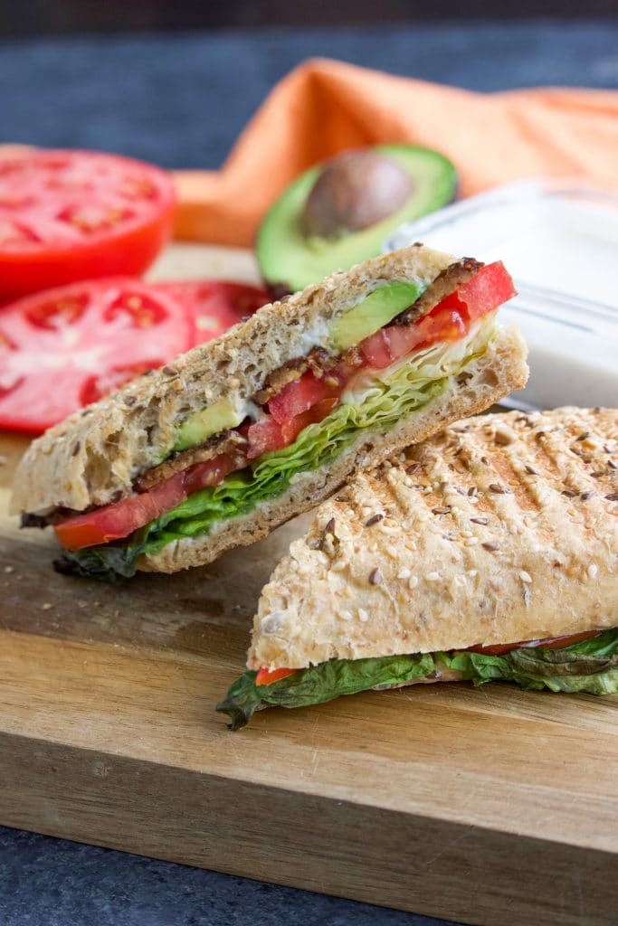 A TTLA Whole Foods vegan sandwich cut in half and stacked on each other at an angle with a tomato and avocado behind it on top of a cutting board. 