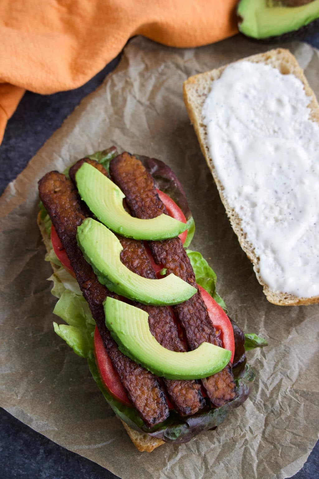 A Whole Foods TTLA sandwich with the top roll next to it on a crinkly piece of parchment paper. 