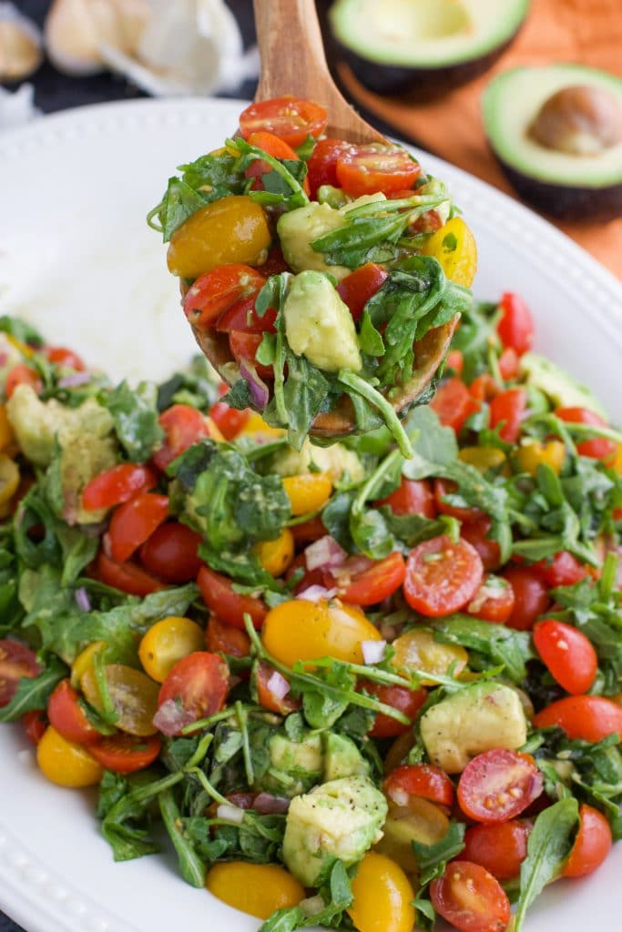Heirloom Cherry Tomato, Avocado and Arugula Salad tossed together on a white plate and served with a wooden spoon