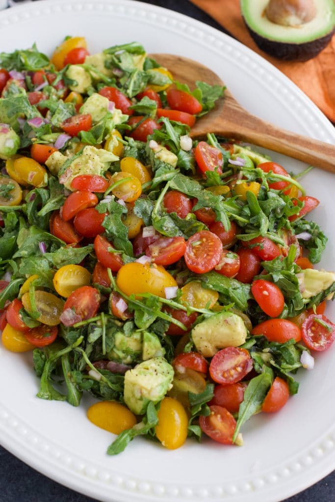 A large white platter filled with vegan arugula salad and a wooden spoon next to an orange napkin.