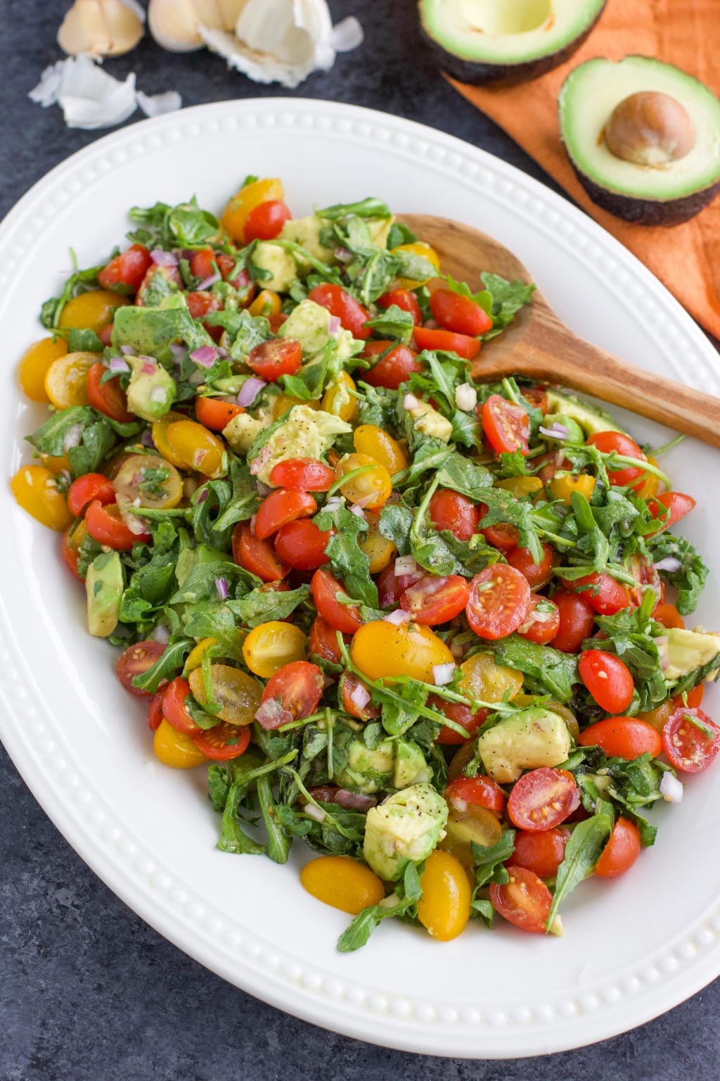 Low-carb vegan tomato salad with avocado, arugula, red onion, and balsamic vinaigrette on a large white platter with a wooden spoon. 