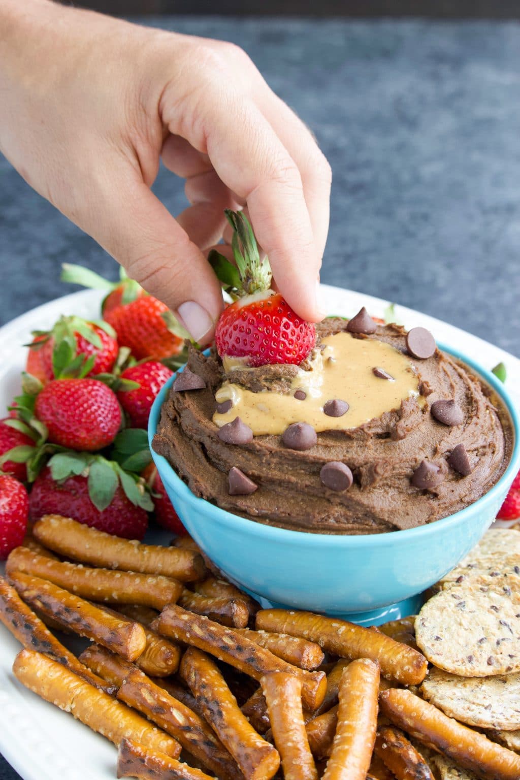 A hand dipping a strawberry into a bowl of dessert hummus on a platter with snacks. 